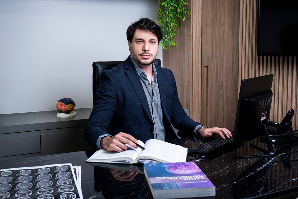 Dr. Rossini Monteiro, neurologista, sentado à mesa do consultório, com um livro de neurologia aberto e exames de imagem ao lado.